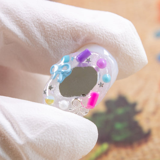Gloved hand holding a clear press-on nail tip decorated with a mini mirror piece, bow, and beads.