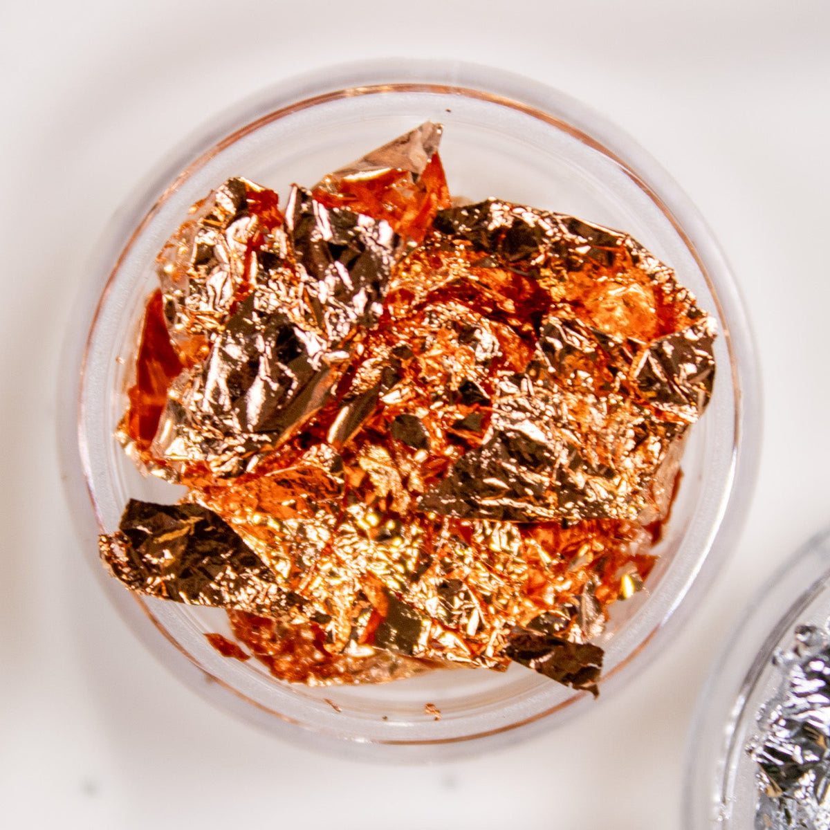 Close-up view of rose gold metallic nail foil flakes in a small clear container.