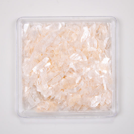 Close-up view of peach nude pearl shell flakes in a small clear square container.
