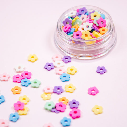 Pastel flower nail charms in pink, purple, yellow, and blue scattered across a pink background.