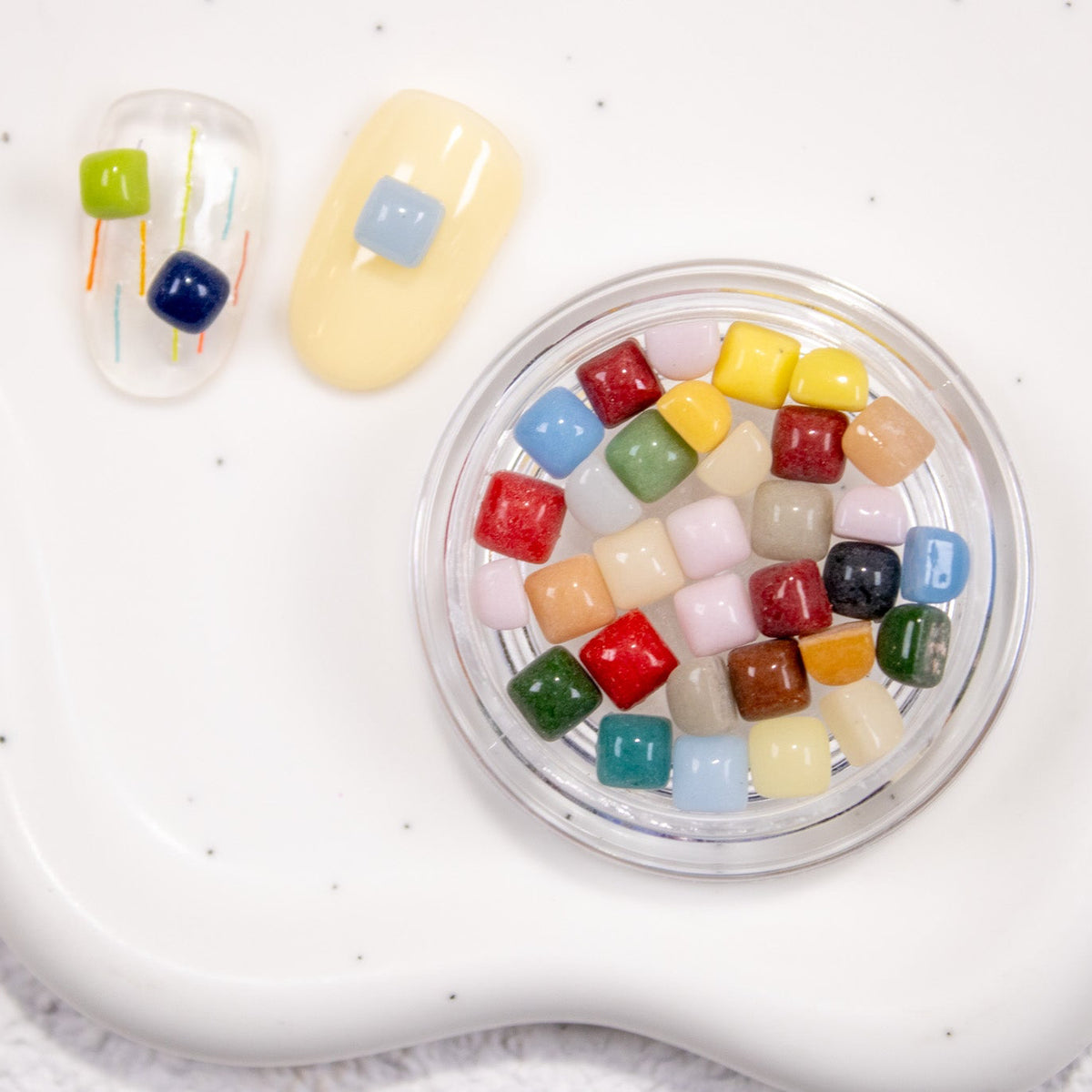 Jar of colorful cube nail stones placed on a white wavy ceramic tray with sample nails.