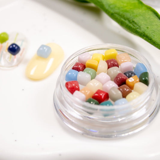 Clear round jar of colorful jelly bean cube stones next to a green leaf on a white background.