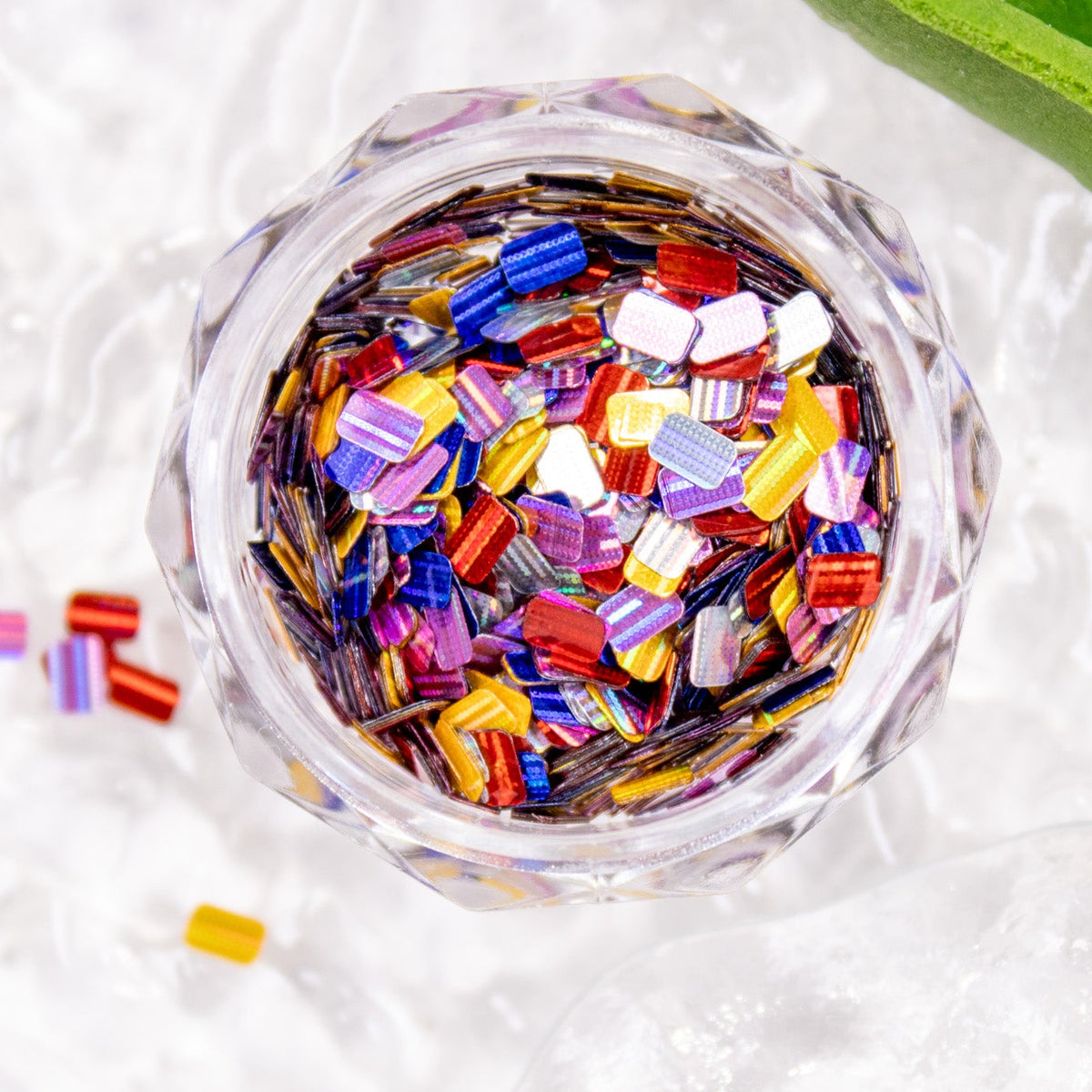 Angled shot of a jar packed with colorful mini bar glitter next to green leaves.