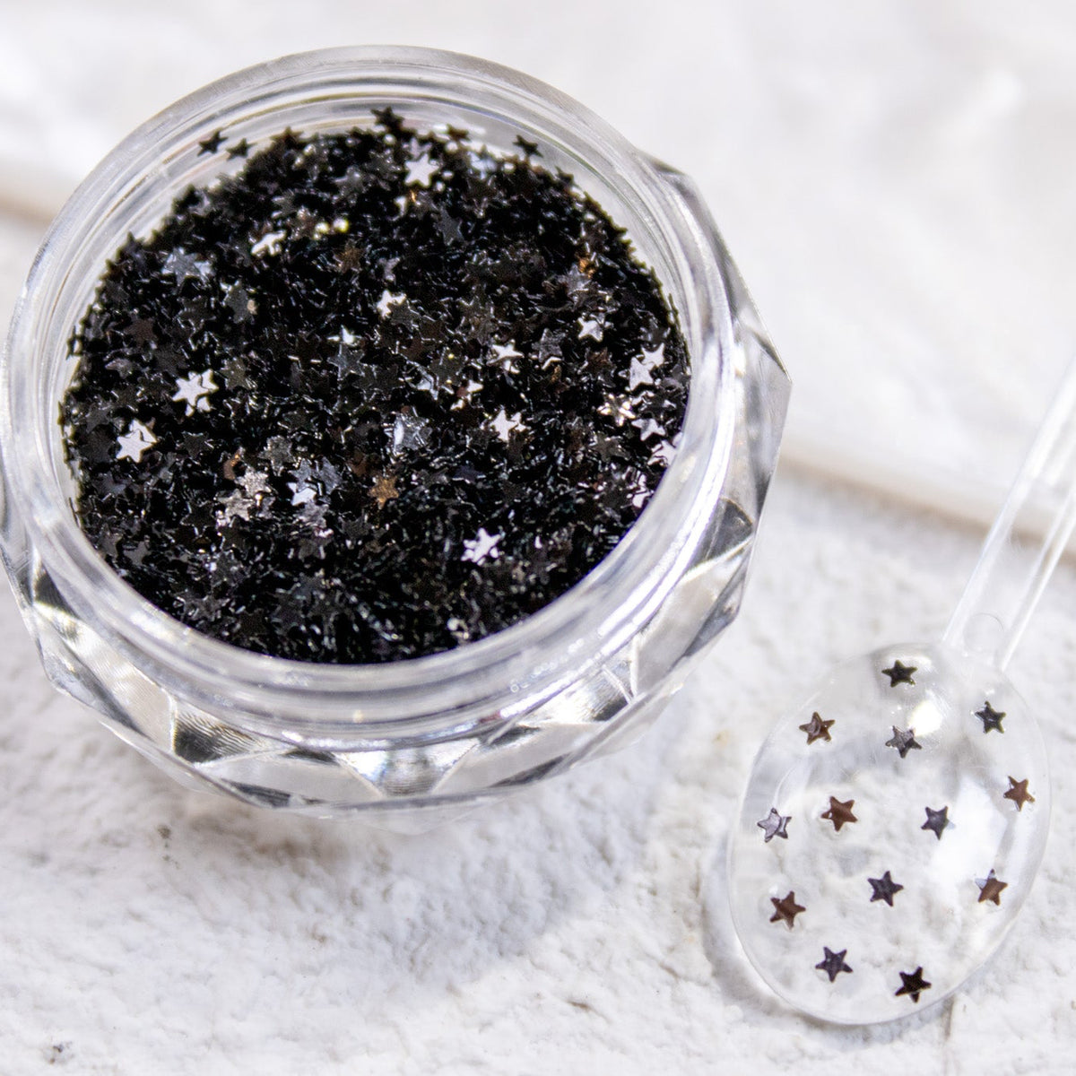 Close-up view of a clear jar filled with black mini star nail glitter.
