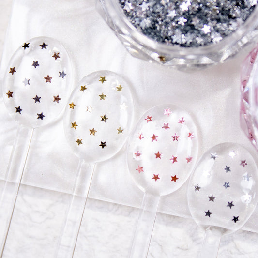 Four small white spoons displaying pink, silver, gold, and black mini star nail glitter samples.