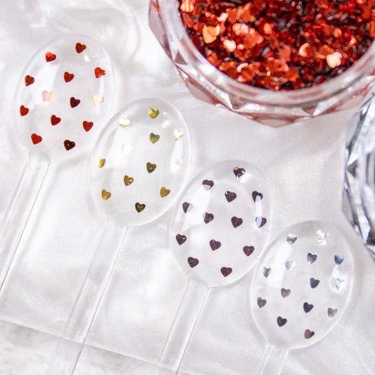 Four small white spoons displaying red, black, silver, and gold mini heart nail glitter samples.
