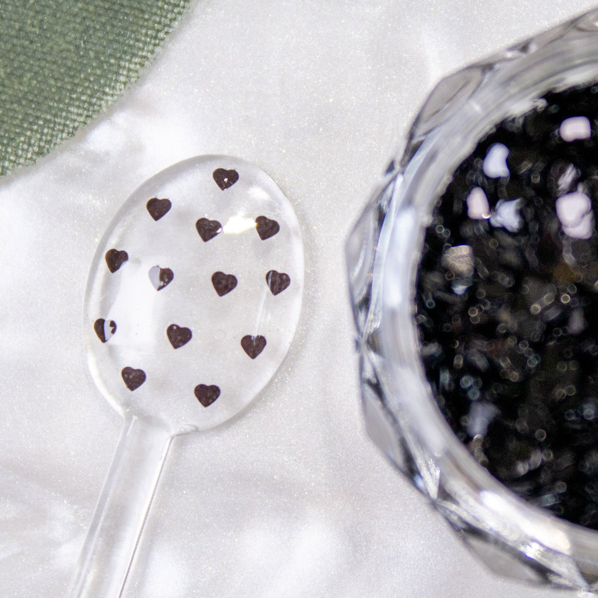 Close-up view of a clear jar filled with black mini heart nail glitter next to a spoon sample.