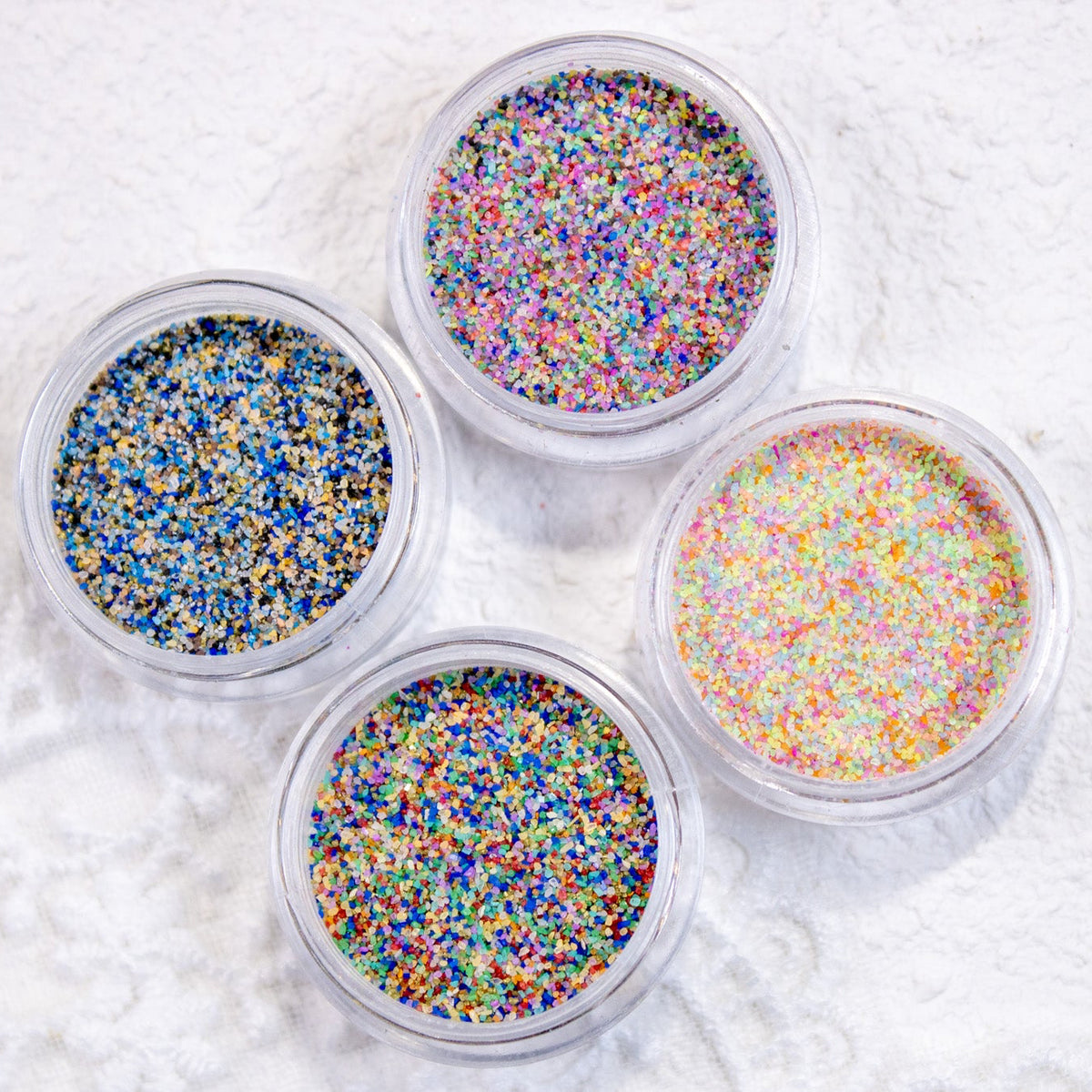 Top-down flat lay of four clear jars displaying fine sugar powder glitter mixes on white lace fabric.
