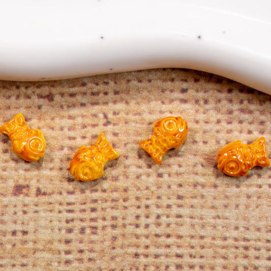 Four miniature orange bungeoppang fish pastry nail charms placed on a textured burlap cloth.