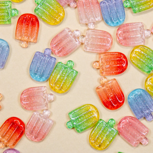 Top-down flat lay of mixed color glitter mini popsicle 3D nail charms spread across a light background.