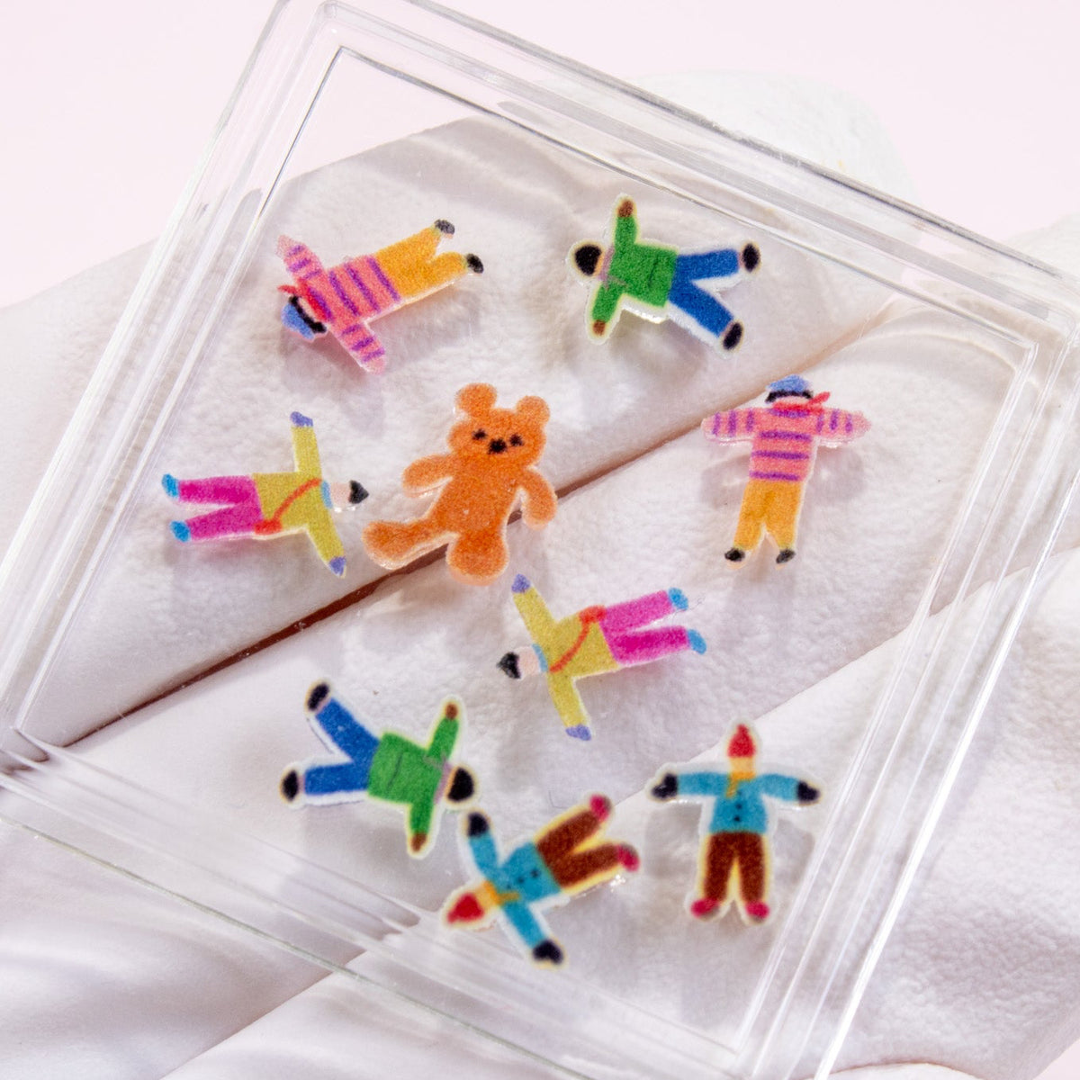 Gloved hand holding a clear case filled with tiny colorful people and teddy bear 3D nail charms.