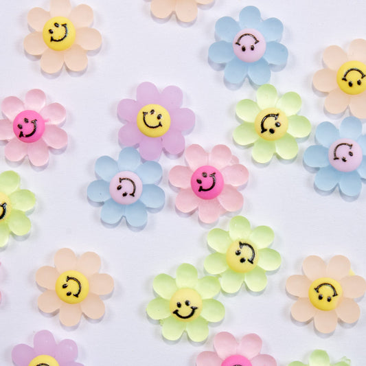 Top-down flat lay pattern of pastel smiley-face daisy 3D nail charms in pink, orange, yellow, green, blue, and purple.