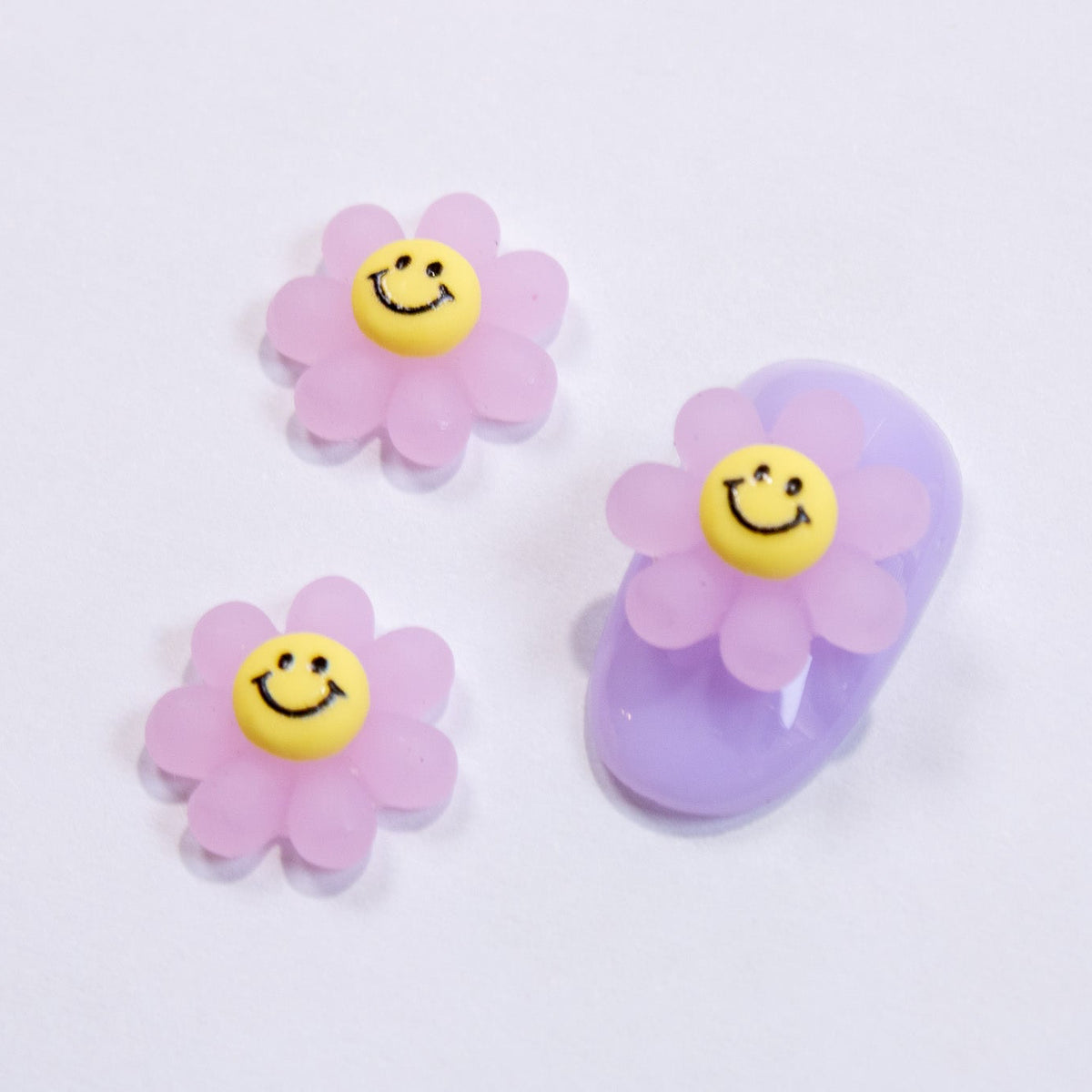 Pastel purple press-on nail swatch paired with two purple smiley flower 3D nail charms.