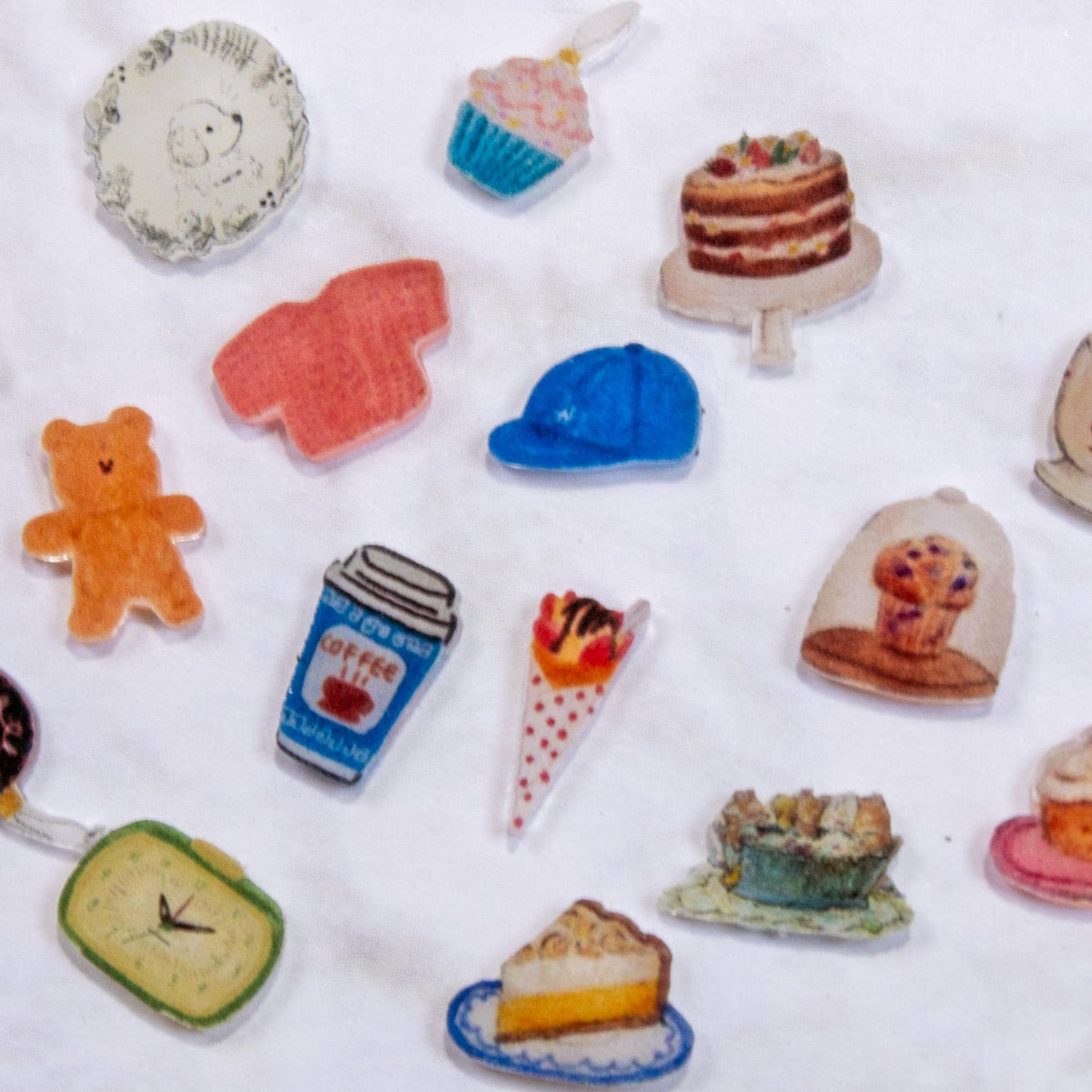 Top-down view of assorted miniature afternoon tea charms including cakes, pie slices, coffee cups, clocks, utensils, and teddy bears.