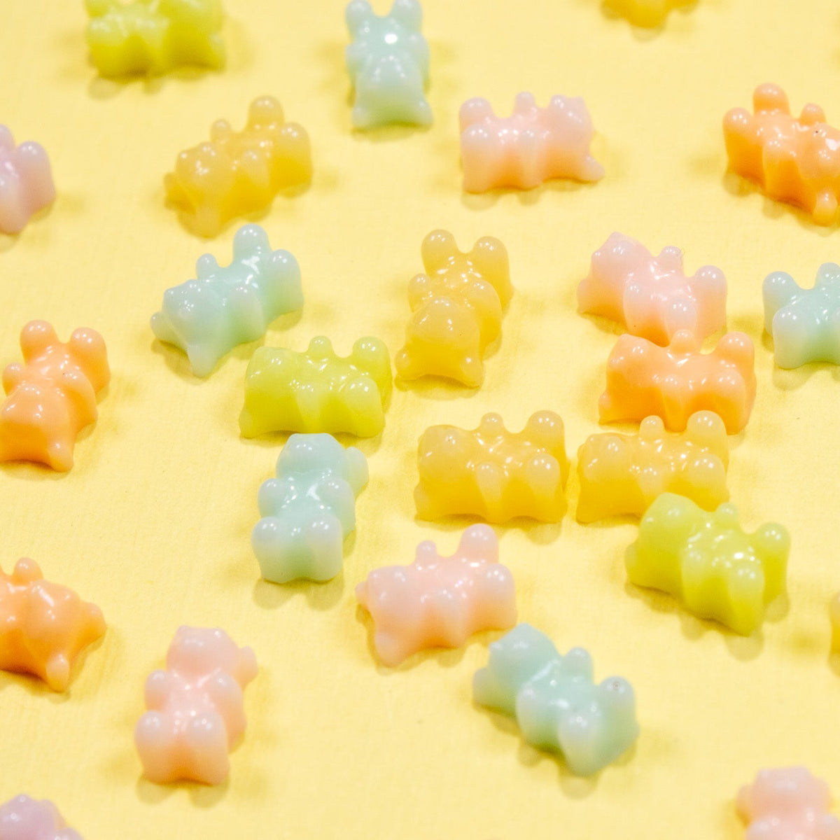 Scattered pattern of colorful miniature pastel gummy bear 3D nail charms spread across a soft yellow background.