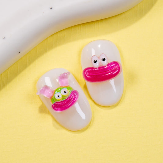 White press-on nail swatches decorated with a wide-eyed cartoon face charm and a pouty pink lips charm on a yellow surface.