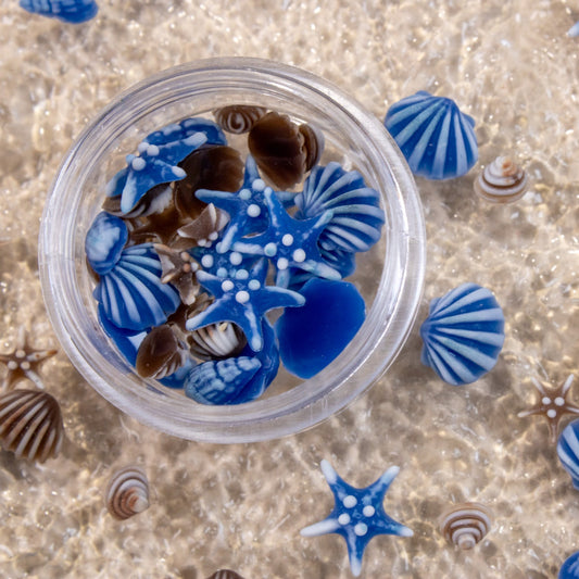 Clear round storage jar filled with mini blue and brown starfish and striped seashell 3D nail charms on brown background.