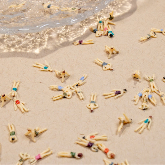 Wide top-down view of miniature swimmer 3D nail charms in colorful bathing suits scattered across a sand-textured surface.