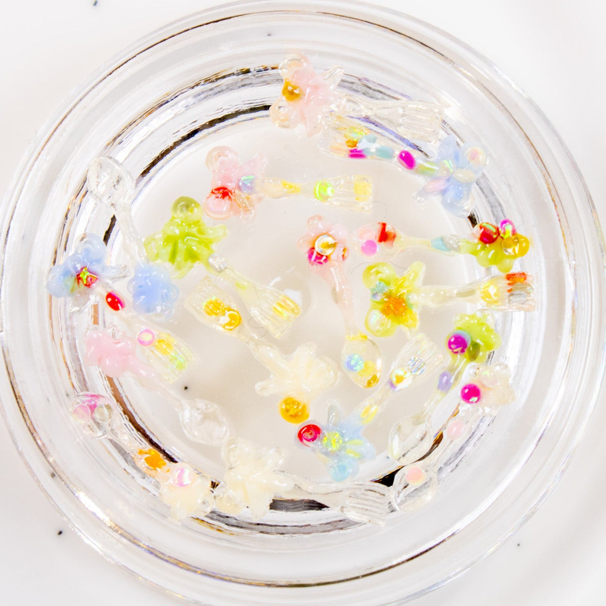 Top-down close-up of a clear jar filled with colorful mini utensil 3D nail charms on a ceramic dish.