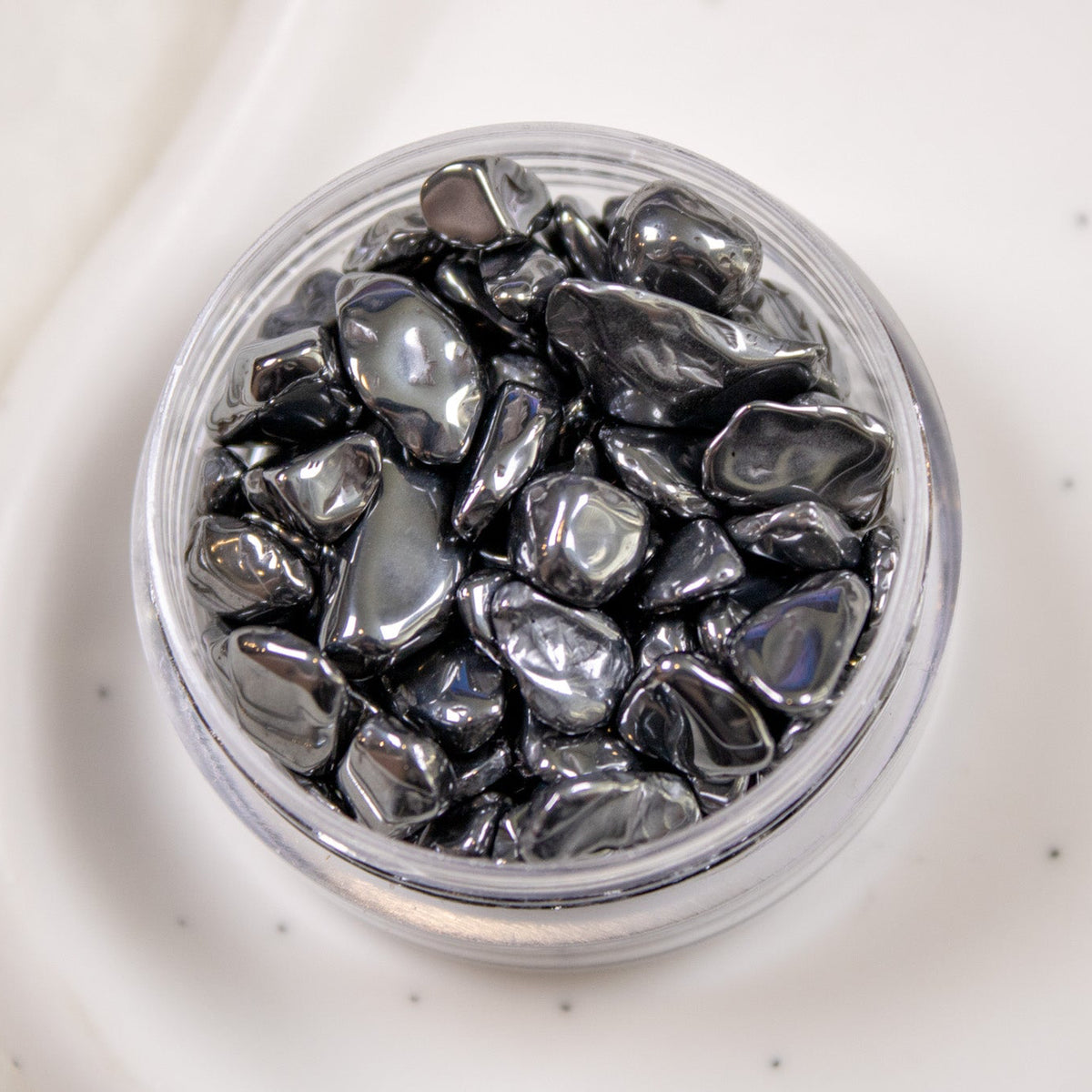 Top-down view of an open clear mini jar completely filled with glossy metallic black hematite-style nail stones.