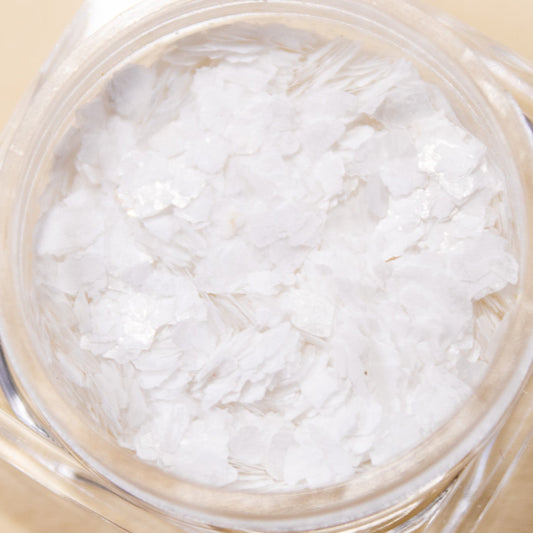 Close-up top-down view of an open jar filled with irregular white snowflake nail glitter flakes.