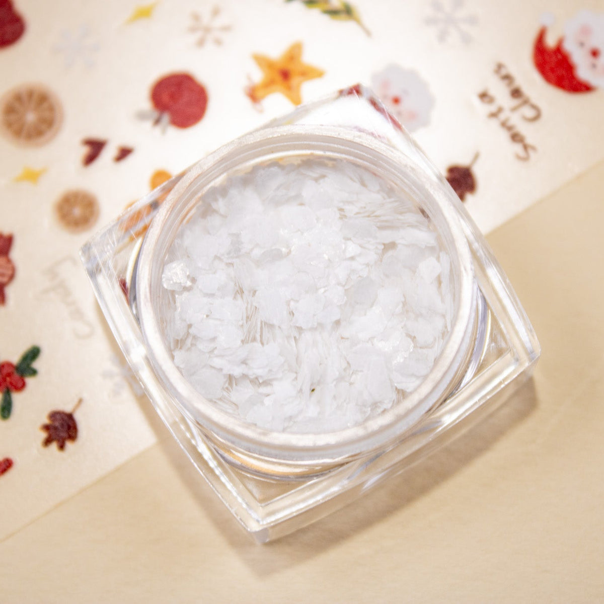 Angled view of an open jar of Mini Snowflake Glitter on a Christmas-themed background.
