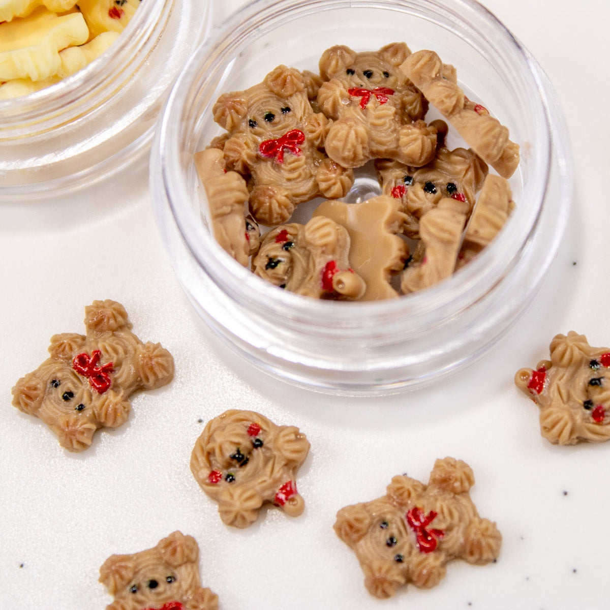 Chocolate cookie bear nail charms with tiny red bow accents arranged on a white surface with a jar.