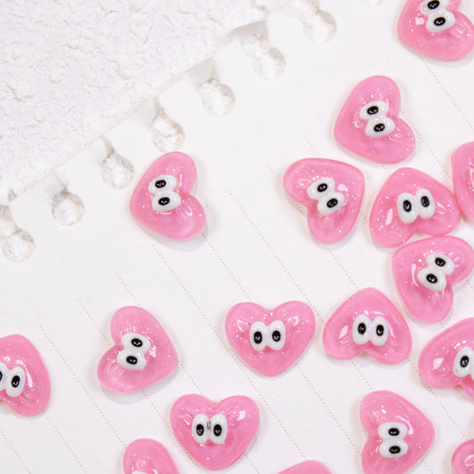 Top-down flat lay pattern of glossy pink heart 3D nail charms with googly cartoon eyes.