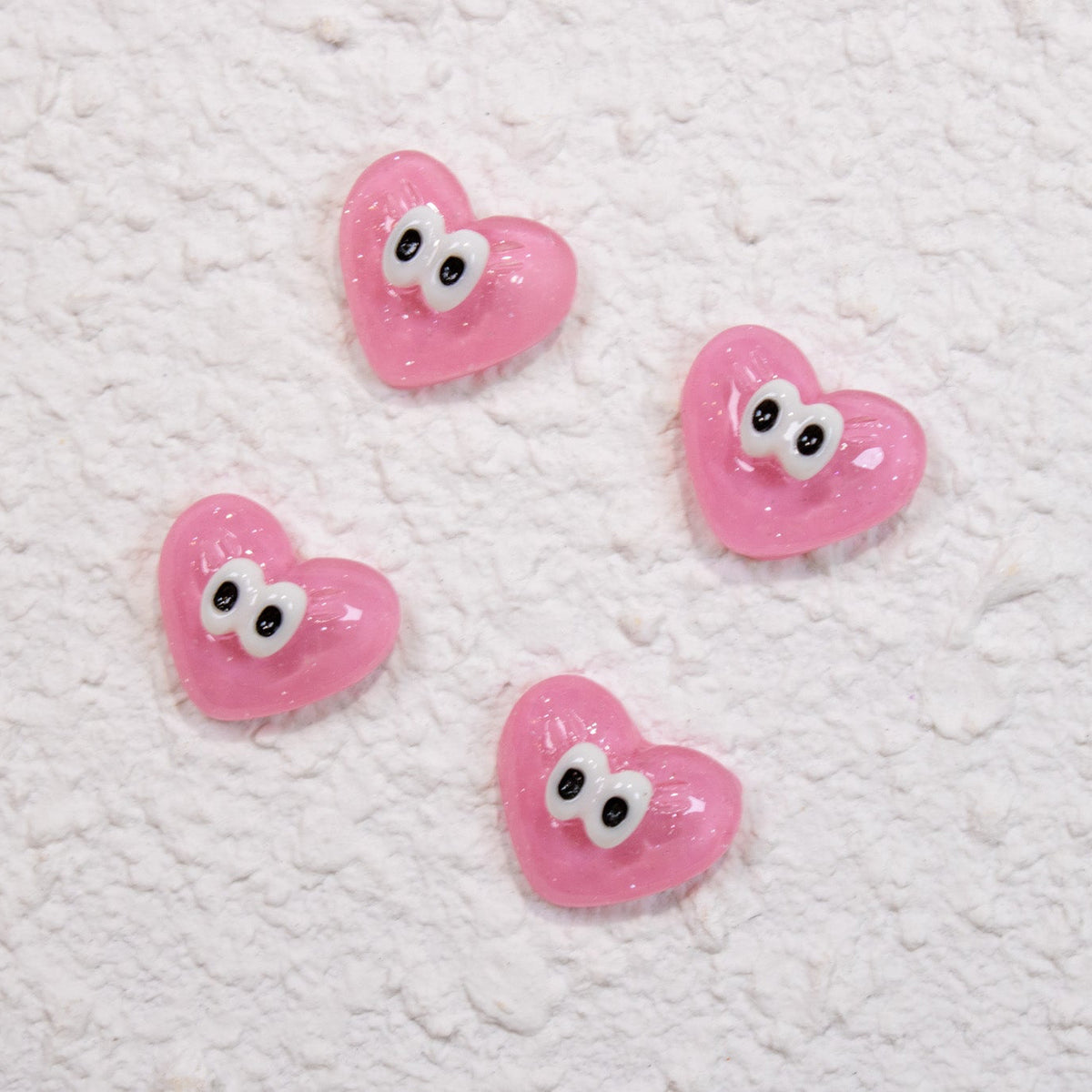 Four glossy pink googly-eye heart 3D nail charms displayed on a textured white surface.