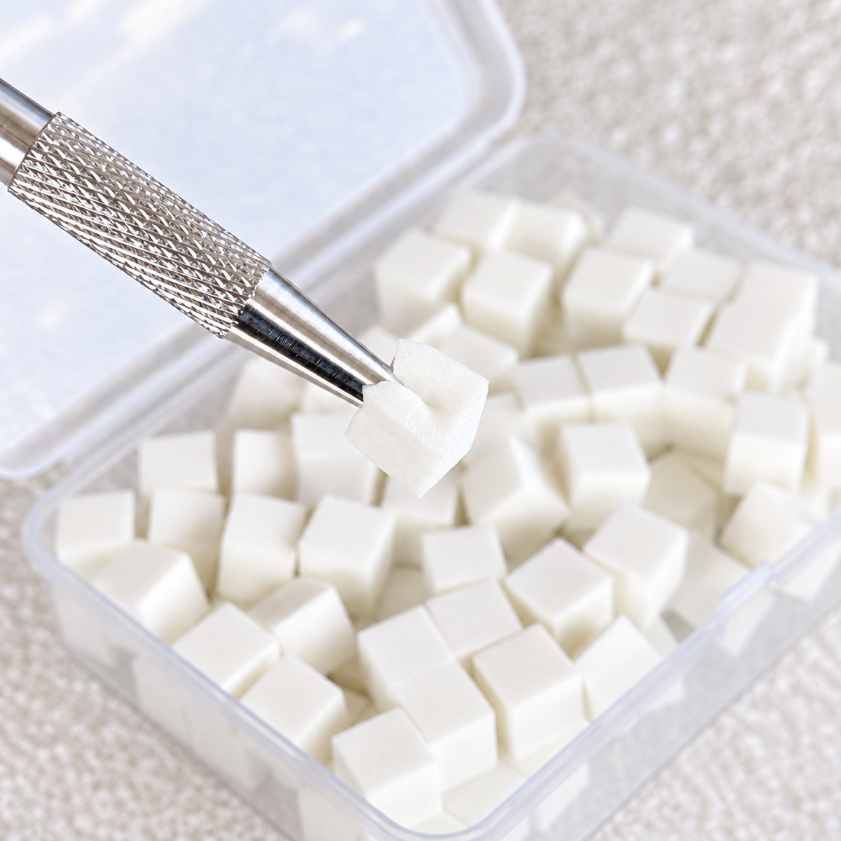 Precision metal grabber tool picking up a single white mini nail art sponge from the clear storage case.