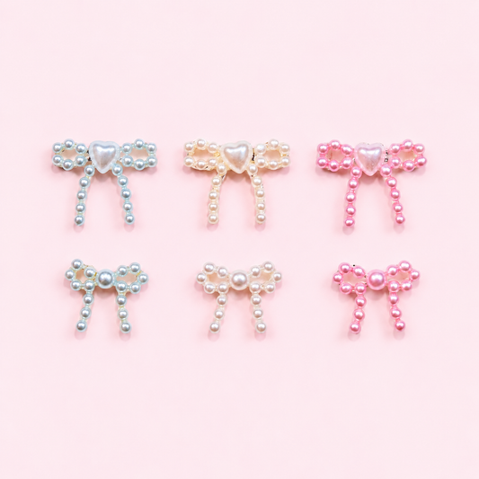 Three large and three small pearl ribbon bow 3D nail charms with heart or center accents in blue, white, and pink arranged in two rows on a soft pink background.