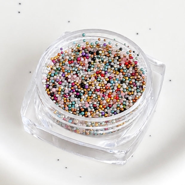 Angled close-up of an open square jar filled with assorted multicolor metallic micro caviar beads.