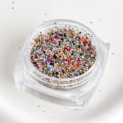 Angled close-up of an open square jar filled with assorted multicolor metallic micro caviar beads.