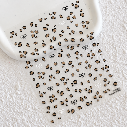 Full sheet of leopard print nail stickers featuring classic brown leopard spots and black and white mini bow accents on a scalloped ceramic tray.