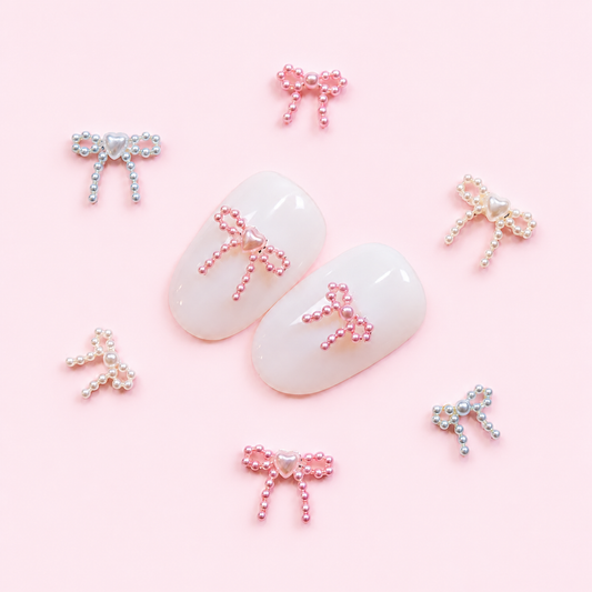 White press-on nail swatches displaying pink pearl ribbon bow 3D nail charms surrounded by scattered pink, white, and blue pearl bows on a soft pink backdrop.