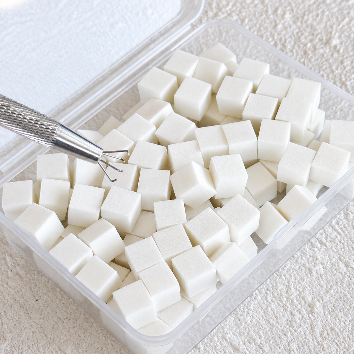 Close-up shot of the silver metal grabber pen positioned over the white mini nail sponges inside the clear plastic case.
