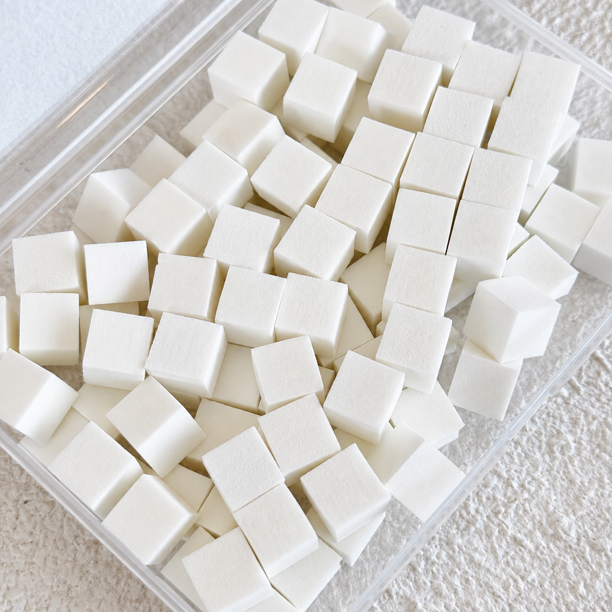 Top-down view of a clear plastic container packed with 100 small white cube-shaped mini nail art sponges.