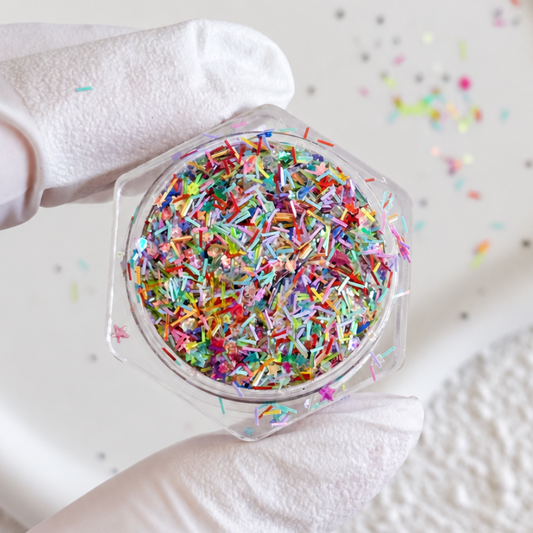 Gloved hand holding an open jar filled with a colorful mix of sprinkle-like glitter strands and tiny star sequins.