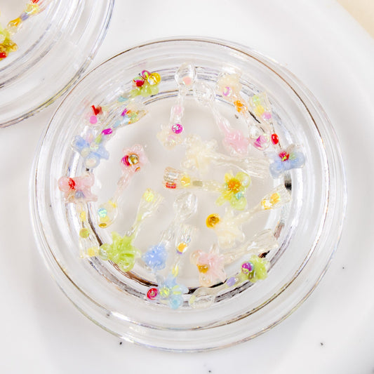 Two clear round jars filled with colorful mini spoon and fork 3D nail charms resting on a white speckled plate.